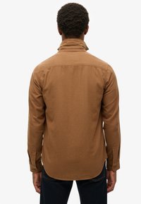 Bruine langearmige shirt met een relaxte pasvorm, hoge kraag, knoopmanchetten en details op de rug. Zachte textuur.