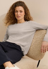 Pullover grigio chiaro in maglia con scollatura a girocollo e polsini a coste, abbinato a pantaloni blu scuro a trama. Seduto su una morbida poltrona beige.