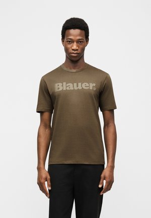 Mladić u maslinasto zelenoj majici s natpisom "Blauer" i crnim hlačama, stoji ispred jednobojne svijetle pozadine.