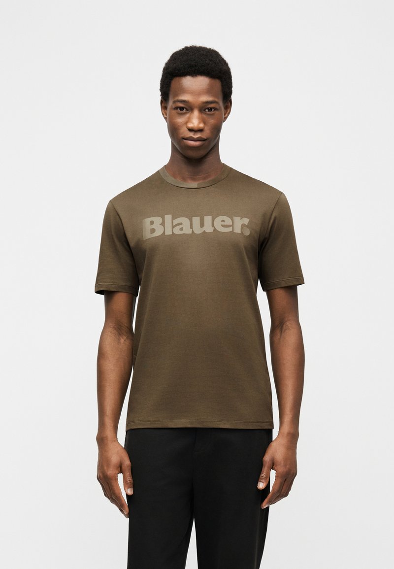 Νεαρός άνδρας που φοράει πράσινο ελιάς μπλουζάκι με το κείμενο "Blauer" και μαύρο παντελόνι, στέκεται μπροστά σε ένα απλό φωτεινό φόντο.