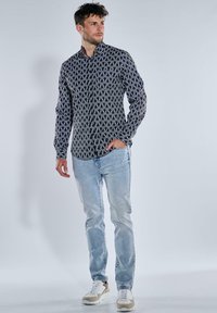 Schwarz-weißer gemusterter Hemd, langärmlig, kombiniert mit hellblauen Jeans und neutral-tonigen Sneakers.