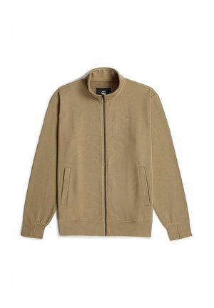 Chaqueta beige con cremallera, cuello alto, mangas largas, dos bolsillos laterales y una textura suave y lisa. Logotipo minimalista en el pecho.