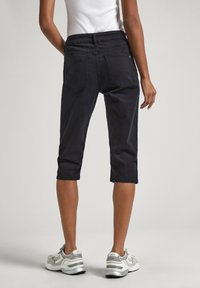 Pantalones cortos negros con una textura suave, con dos bolsillos traseros y una cinturilla de altura media, combinados con zapatillas deportivas grises.