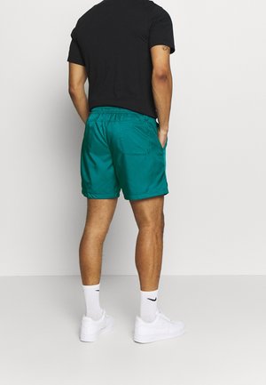 Zielone szorty wykonane z gładkiego materiału z elastycznym paskiem w talii, posiadające tylnią kieszeń i boczne szwy. Noszone z czarną koszulką i białymi trampkami.