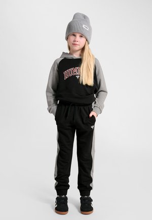 Barn med langt blond hår iført grå strikket hue, sort og grå hættetrøje med teksten "HUMMEL", matchende bukser og sorte sneakers, står med hænderne i lommerne.