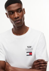 Νέος άντρας που φοράει λευκό μπλουζάκι με τα λογότυπα της ομάδας Formula 1 της Cadillac και της Tommy Hilfiger, με τα χέρια σταυρωμένα, κοιτάζει ελαφρώς προς το πλάι.