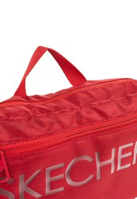 Bolso de tela roja con un acabado texturizado, que cuenta con un zipper superior y una asa resistente, marcado con la palabra "SKECHERS" en letras blancas.