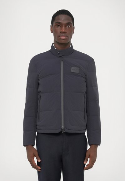 Dunkelblaue Puffjacke mit hohem Kragen, Frontreißverschluss und horizontalem Steppmuster. Mit seitlichen Taschen und einem Logopatch.