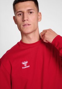 Czerwony długi rękaw crewneck z miękką teksturą. Posiada białe haftowane logo na klatce piersiowej. Prosty, nowoczesny design z luźnym krojem.
