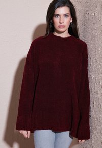 Burgundy oversized knit sweater with a textured finish, crew neck, and wide sleeves. Paired with light blue jeans.