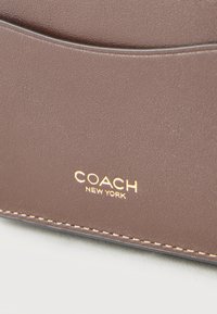 Καφέ δερμάτινη επιφάνεια με χρυσό λογότυπο "COACH NEW YORK" και μπεζ ραφές κατά μήκος της άκρης μιας τσάντας ή πορτοφολιού.