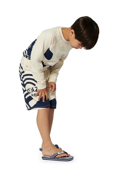 Kind in een crèmekleurig sweatshirt met marineblauwe accenten en gebloemde shorts. Marineblauwe slippers maken de outfit af. Staande en kijkt naar zijn been.