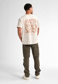 T-shirt en coton couleur crème arborant un grand motif floral aux teintes terreuses au dos, associé à un pantalon cargo olive et des baskets blanches.
