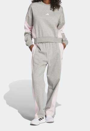 Femme portant un survêtement gris et rose clair, mains dans les poches et baskets blanches avec bandes noires sur fond uni.