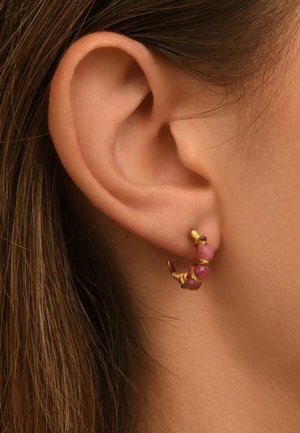 Boucle d'oreille dorée présentant un design courbé avec trois gemstones roses, enveloppées dans un fil délicat, sur un fond de teinte peau.