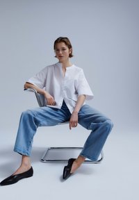 Young person sitting on a modern metal chair, wearing a white loose shirt, blue jeans, and black pointed flats.