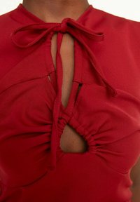 Rode top met een striksluiting en geplooide details rond een centraal uitsnijding. Gemaakt van zachte stof, het heeft een gladde textuur en een strak ontwerp.