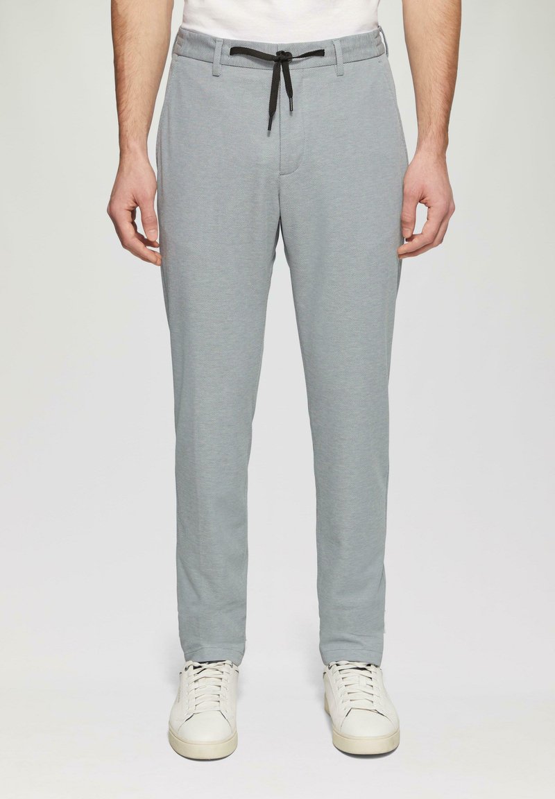 Helle graue Hose mit strukturierten Oberflächen, Kordelzugbund und Seitentaschen, kombiniert mit weißen Sneakers.