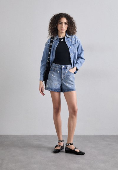 Giacca di jeans con decorazioni di perle, top nero e pantaloni a bermuda blu decorati con fiori; ballerine nere con cinturini borchiati.