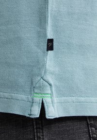 Světle modrá košile s límečkem a texturovanou tkaninou, s zeleným výšivkovým akcentem a černou etiketou s logem na bočním švu.