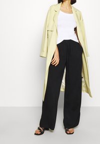 Světle žlutý trenčkot přes bílý tílko, doplněný volnými černými kalhotami a černými sandály na pásky.