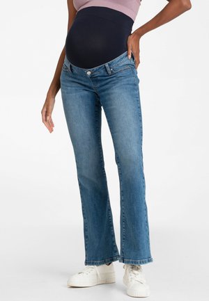 Gravid person iført blå graviditetsjeans med elastisk sort linning og hvide sneakers stående mod en hvid baggrund.