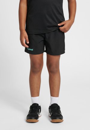 Dziecko noszące czarne spodenki sportowe z logo "hummel", czarno-białe buty sportowe, białe skarpetki i czarny top ciągnący za brzeg.