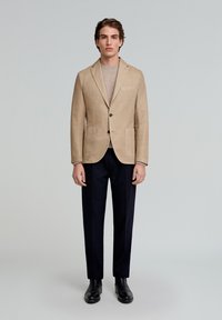 Blazer de lino beige con solapas de muesca, cierre de un solo botón y dos bolsillos delanteros, combinado con un suéter claro y pantalones oscuros.