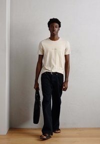 Tricou din bumbac bej cu un grafic subtil, blugi negri din denim, geantă de umăr neagră și șlapi maro, stând cu spatele la un perete deschis.