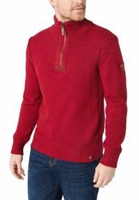 Roter Strickpullover mit hohem Kragen, halber Reißverschluss, gerippten Bündchen und Saum sowie einem kleinen Logo-Patch am linken Ärmel.