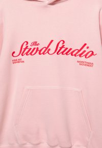 Világos rózsaszín pulóver elülső kenguruzsebbel. Piros hímzett szöveget tartalmaz, amely "The Stvd Studio" és "FINE ART EXHIBITION" feliratokat mutatja vastag betűtípussal.