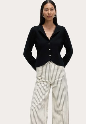 Sort knap-op cardigan med en glat tekstur, der har en sjalskrave og sølvknapper. Matchet med hvide bukser med brede ben og striber.