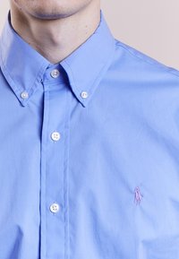 Ljusblå skjorta med knappar i bomull. Har vita knappar, en spetskrage och en liten broderad logotyp på vänster bröst.