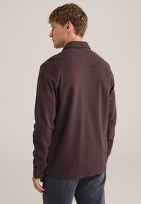 Rjava dolgosleeve srajca iz teksturirane tkanine s ovratnikom, ki ima zadnji šiv in gumbne detajle na manšetah.