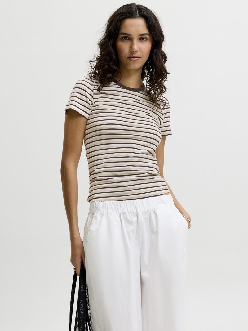 Femme aux cheveux bouclés et foncés portant un t-shirt rayé marron et beige et un pantalon blanc, tenant un sac noir devant un fond uni.