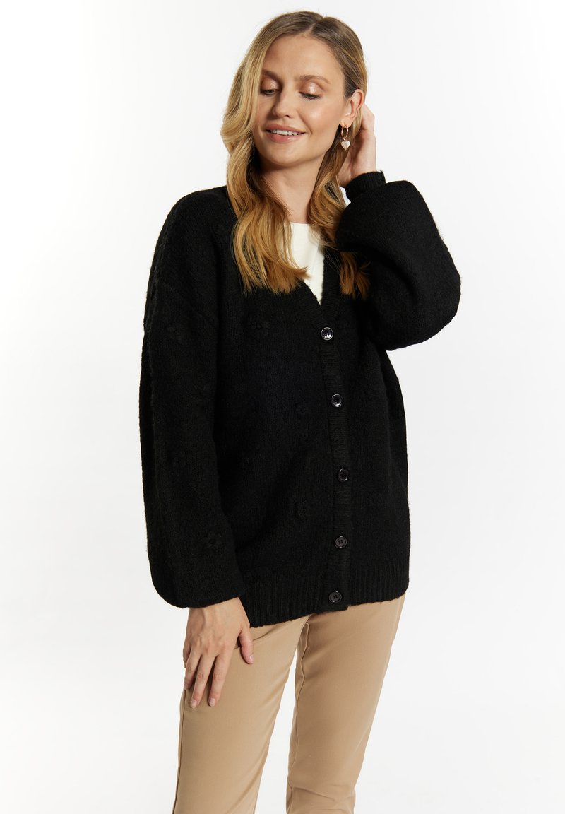 Zwarte cardigan met textuur gebreide patroon, v-hals, oversized silhouet en vijfknops sluiting, gecombineerd met lichtbeige broeken.