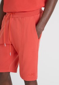 Bermudas deportivas de color coral, fabricadas con material suave, que cuentan con una cintura con cordón, bolsillos laterales y un logo bordado cerca del dobladillo.