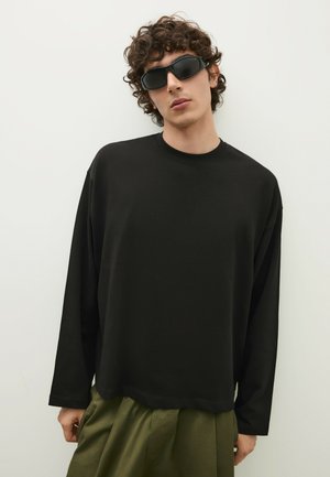 Junge Person mit lockigem Haar, trägt ein schwarzes übergroßes Langarmshirt, olivgrüne Hose und dunkle Rundumsonnenbrille vor weißem Hintergrund.