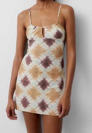 Femme portant une mini robe sans manches tie-dye avec de fines bretelles et une petite ouverture en forme de trou de serrure à l'avant, debout devant un fond uni.