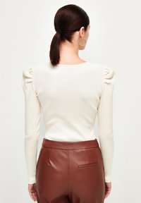 Femme aux cheveux foncés attachés en queue-de-cheval basse, portant un haut crème à manches longues avec des épaules bouffantes et un pantalon en cuir marron taille haute, vue de dos.