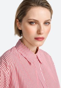 Maglietta a righe rosse e bianche con colletto classico, caratterizzata da una chiusura frontale con bottoni, una trama liscia e motivi verticali uniformi.