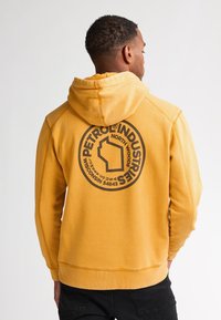 Felpa gialla realizzata in materiale morbido, con un grande logo rotondo nero con testo sul retro. Cappuccio e polsini a coste.