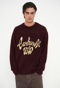 Bordó kötött pulóver, krémszínű logóval, kerek nyakkal és bordás mandzsettával. Puha tapintás, laza fazon és hosszú ujjak.