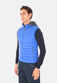 Uomo che indossa un gilet senza maniche trapuntato di un blu vivace sopra una felpa a maniche lunghe scura e jeans blu scuro, in piedi di lato.