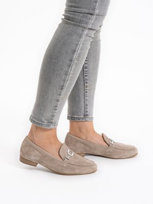 Persona valkā gaiši pelēkas pieguļošas džinsas un bēša zamša loafere ar sudraba sprādzes detaļu, stāvot uz balta fona.