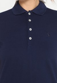 Mörkblå polo-shirt i bomull med krage, fem knappar och en liten broderad logotyp på vänster sida.