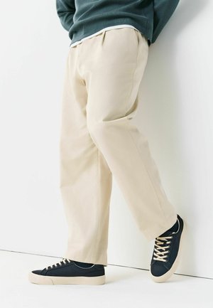 Isik, kes kannab helebeeže värvi pükse ja tumelisi siniseid tossusid kreemikate talladega, seisab valge seina ääres.