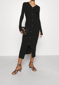 Robe noire en maille côtelée avec manches longues, décolleté en V et fermeture par boutons. Comprend une fente latérale et est associée à des sandales noires à lanières.