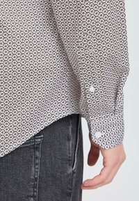 Camisa marrón con pequeño patrón floral blanco, puños abotonados y dobladillo curvado. Llevada con pantalones gris oscuro, mostrando la textura de la tela y el detalle del estampado.