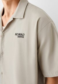 Camisa de manga corta de color beige claro, hecha de tejido suave, con un logo bordado "NOMAD 00456" en negro en el lado izquierdo del pecho.